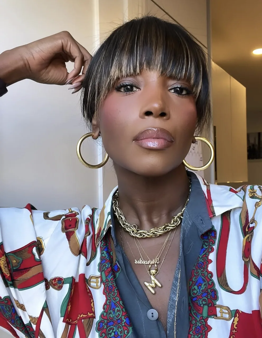 Portrait of Mercedes in thick gold hoops, a gold rope chain with her Mercedes nameplate and an M pendant, and a red-and-white equestrian print blouse over a grey shirt, blunt bangs with highlights