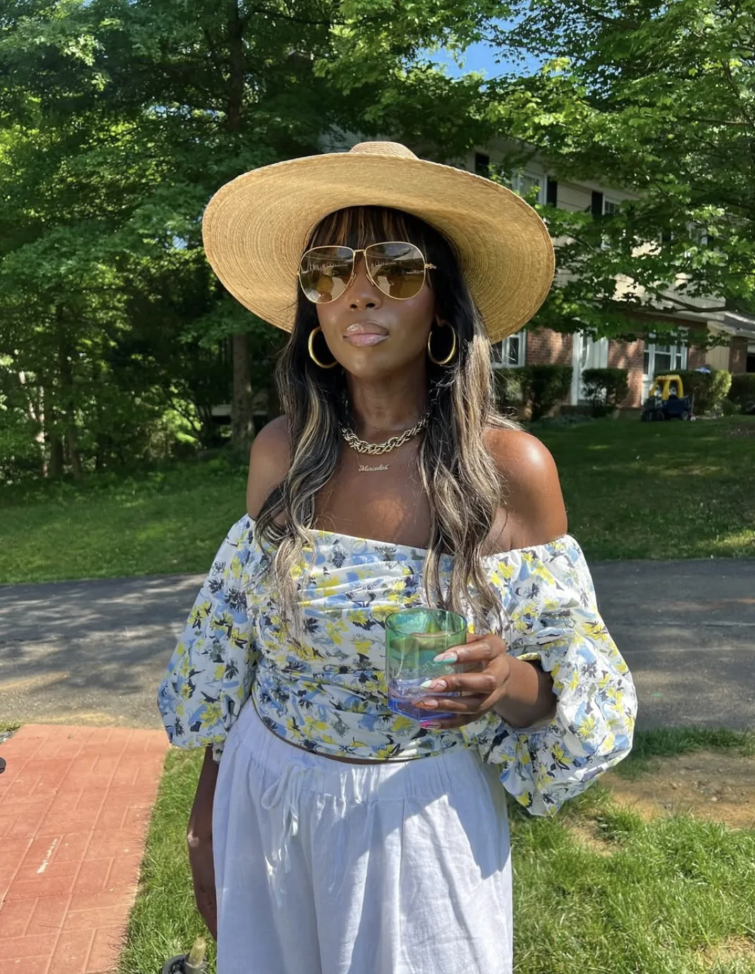 Mercedes on a sunny lawn in a wide straw hat, gold aviators and hoops, an off-shoulder blue and yellow floral top and white linen trousers, holding an iridescent glass