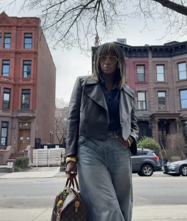 Mercedes on a brownstone-lined street in a cropped black leather jacket, navy shirt, wide light-wash jeans and aviators, carrying a Louis Vuitton bag hung with colorful charms