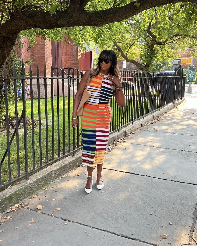 Mercedes on a sunny Brooklyn sidewalk beside an iron fence and a brick church, in a sleeveless multicolor striped midi dress, black sunglasses and white heels